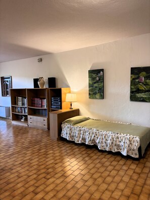 Een kamer met een houten vloer, een bed met bloemenbedekking, een boekenkast, een lamp en twee schilderijen aan de muur.