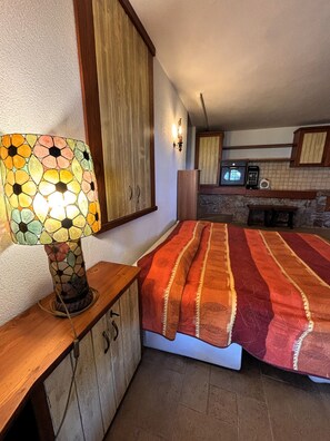A bedroom with a bed, a nightstand, a lamp, a wooden wardrobe, and a kitchen area with a microwave and toaster.