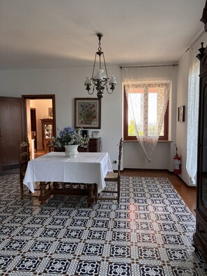 A dining room with a table set for four, a chandelier, patterned floor tiles, and a window with lace curtains.