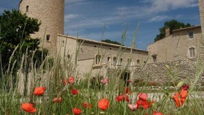 Edificio de piedra con una torre cilíndrica, rodeado de hierba alta y amapolas rojas.