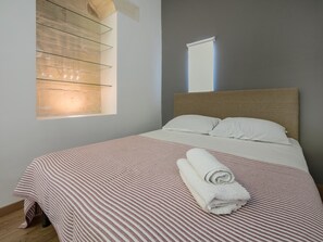 A neatly made bed with a striped bedspread and a rolled towel, a headboard, and a window with blinds.