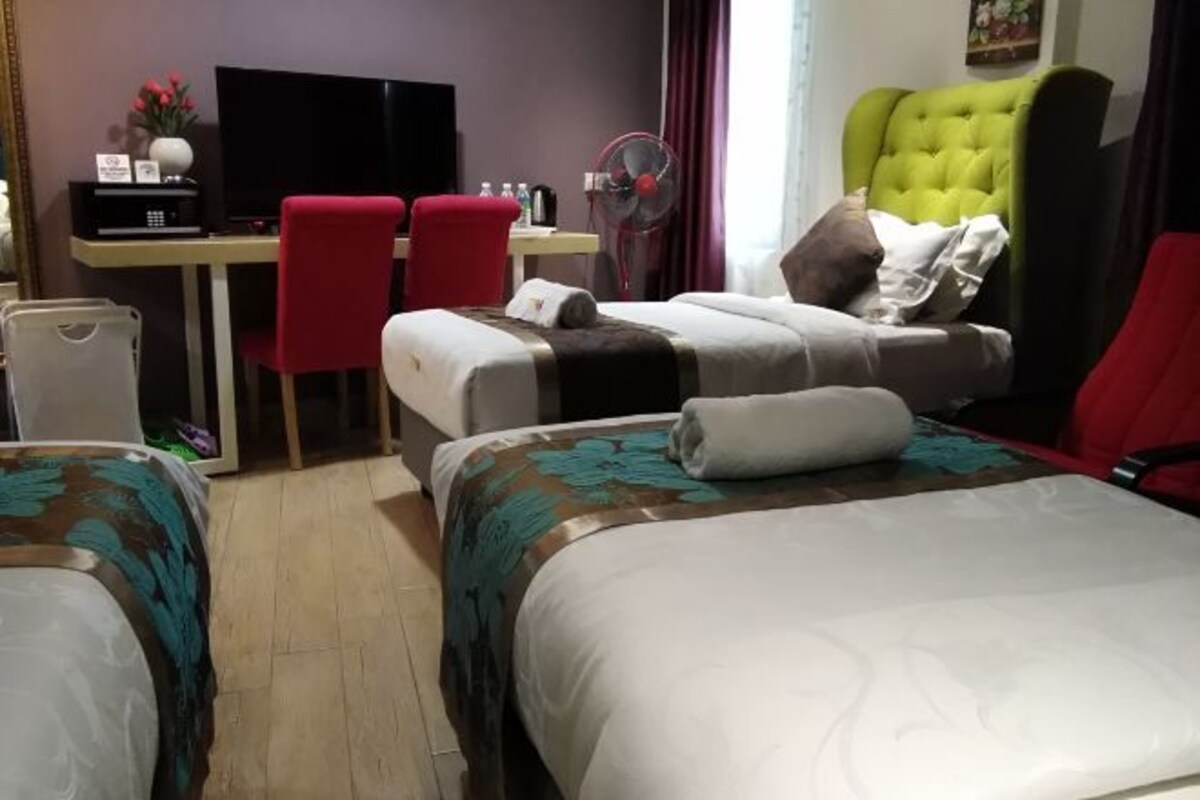 Una habitación de hotel con dos camas, un escritorio con televisor, una silla, un ventilador y un cubo de basura.