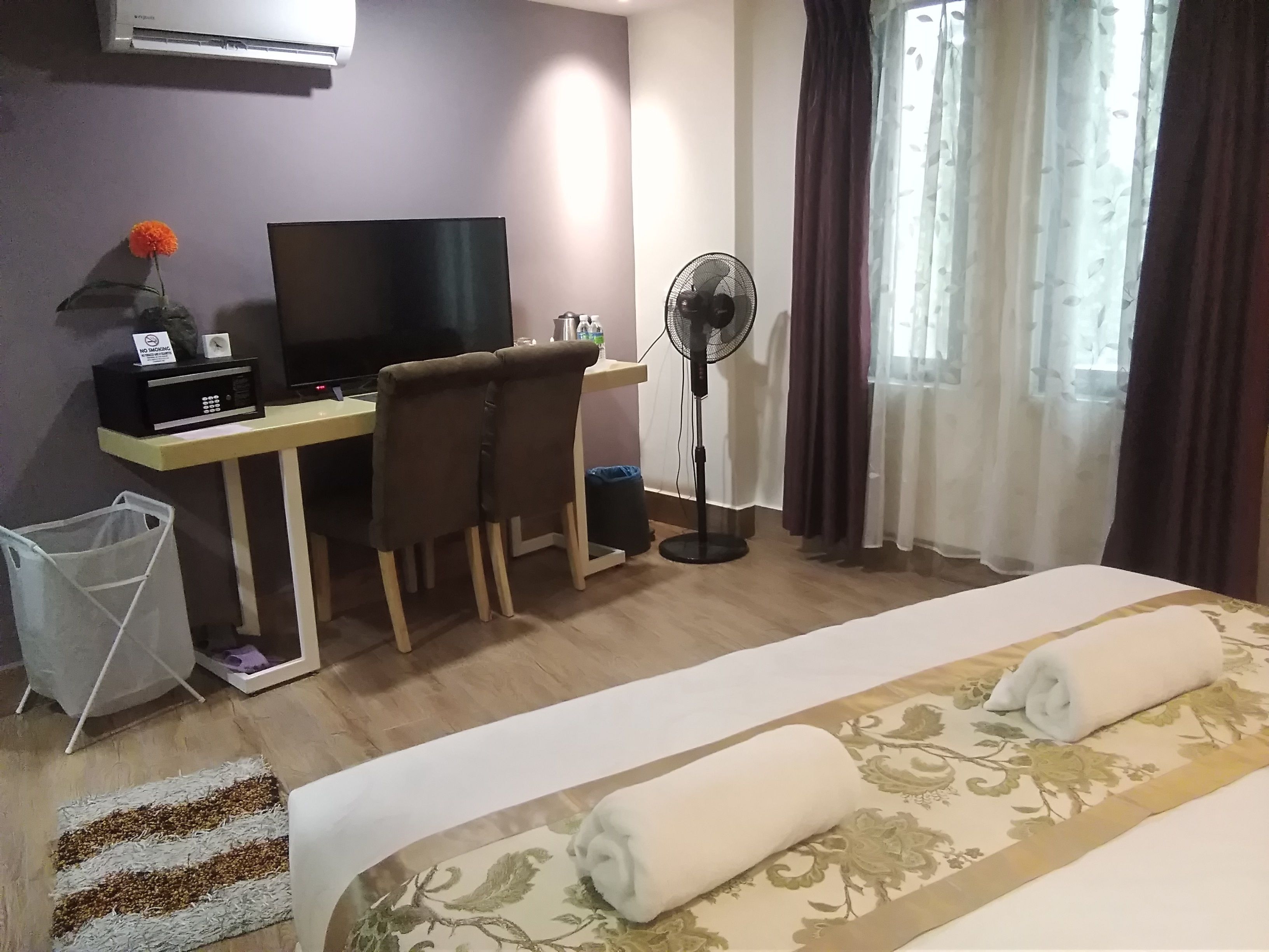 Hotellihuone, jossa on sänky, työpöytä tietokoneella, tuoli, lattiatuuletin ja ikkuna, jossa on verhot.