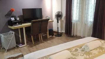 A hotel room with a bed, a desk with a computer, a chair, a standing fan, and a window with curtains.