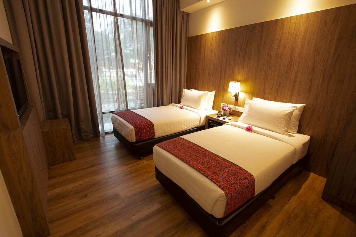 Habitación de hotel con dos camas, piso de madera, un ventanal grande con cortinas y una mesita de noche con lámpara.