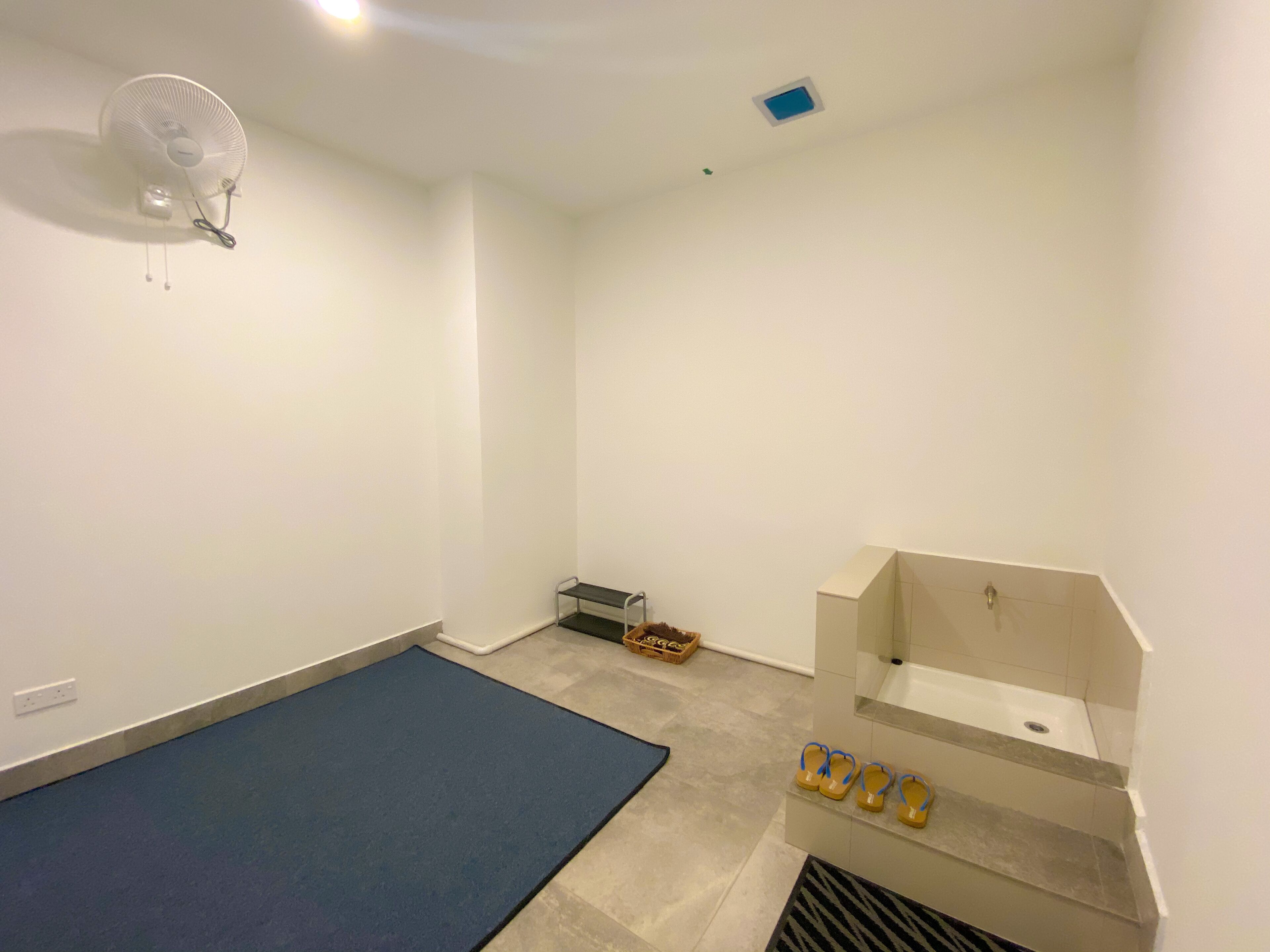 Une petite pièce propre, avec un tapis bleu, un lavabo blanc et un ventilateur au plafond.