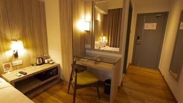Habitación de hotel con piso de madera, un escritorio con silla, cama, espejo, lámpara, teléfono, hervidor de agua y un cubo de basura.