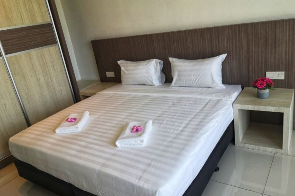A neatly made bed with white linens and two towels with pink bows, a wooden headboard, and a small bedside table with a vase of flowers.