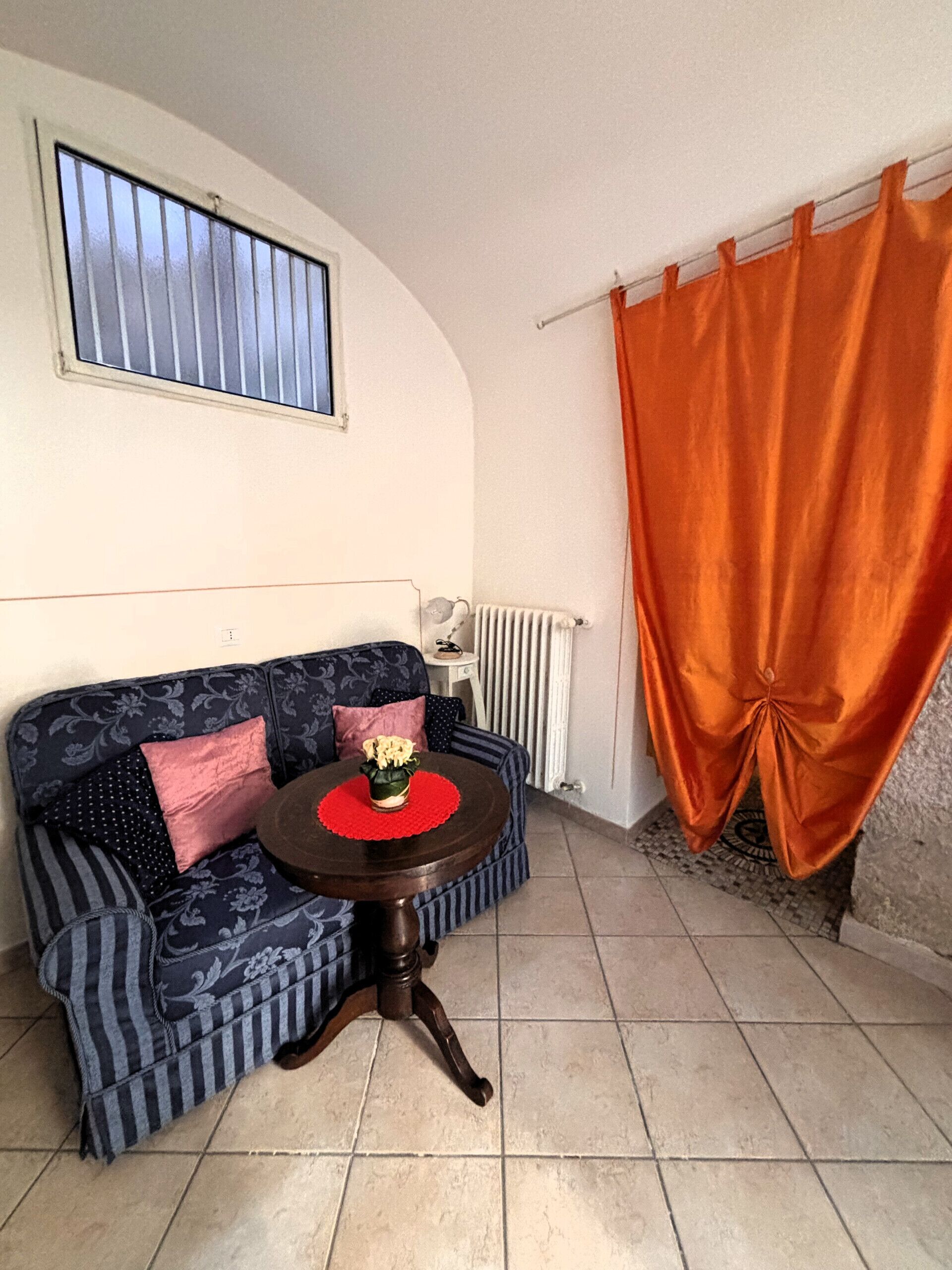 A living room with a sofa, a small round table with a vase, a window with blinds, a radiator, and an orange curtain.