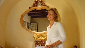 A person sitting on a wooden bench in a room with a large mirror, wooden cabinet, and a decorative wall arch. - 't Aards Paradijs (Deinze)