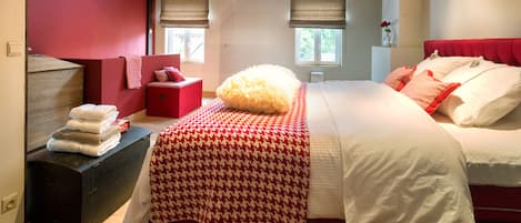Un dormitorio con techo inclinado, cama con ropa de cama blanca y roja, pared de acento roja y ventana con vista a follaje.