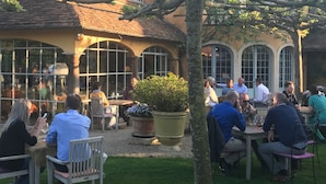 An outdoor patio area with tables and chairs, surrounded by large windows and a tree. - 't Aards Paradijs (Deinze)