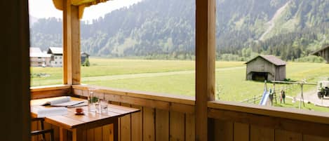 A wooden balcony with a table and chairs, overlooking a green field and a mountain.