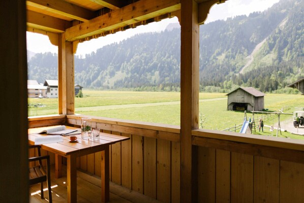 A wooden balcony with a table and chairs, overlooking a green field and a mountain.