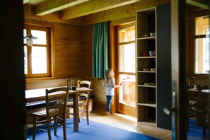 Interior de una cabaña de madera con comedor, un niño cerca de una ventana y estantes con vajilla.