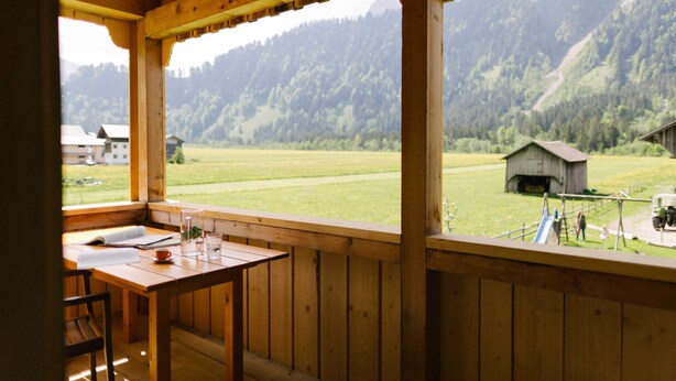 Ein Holzbalkon mit Tisch und Stühlen, der auf ein grünes Feld und einen Berg blickt.