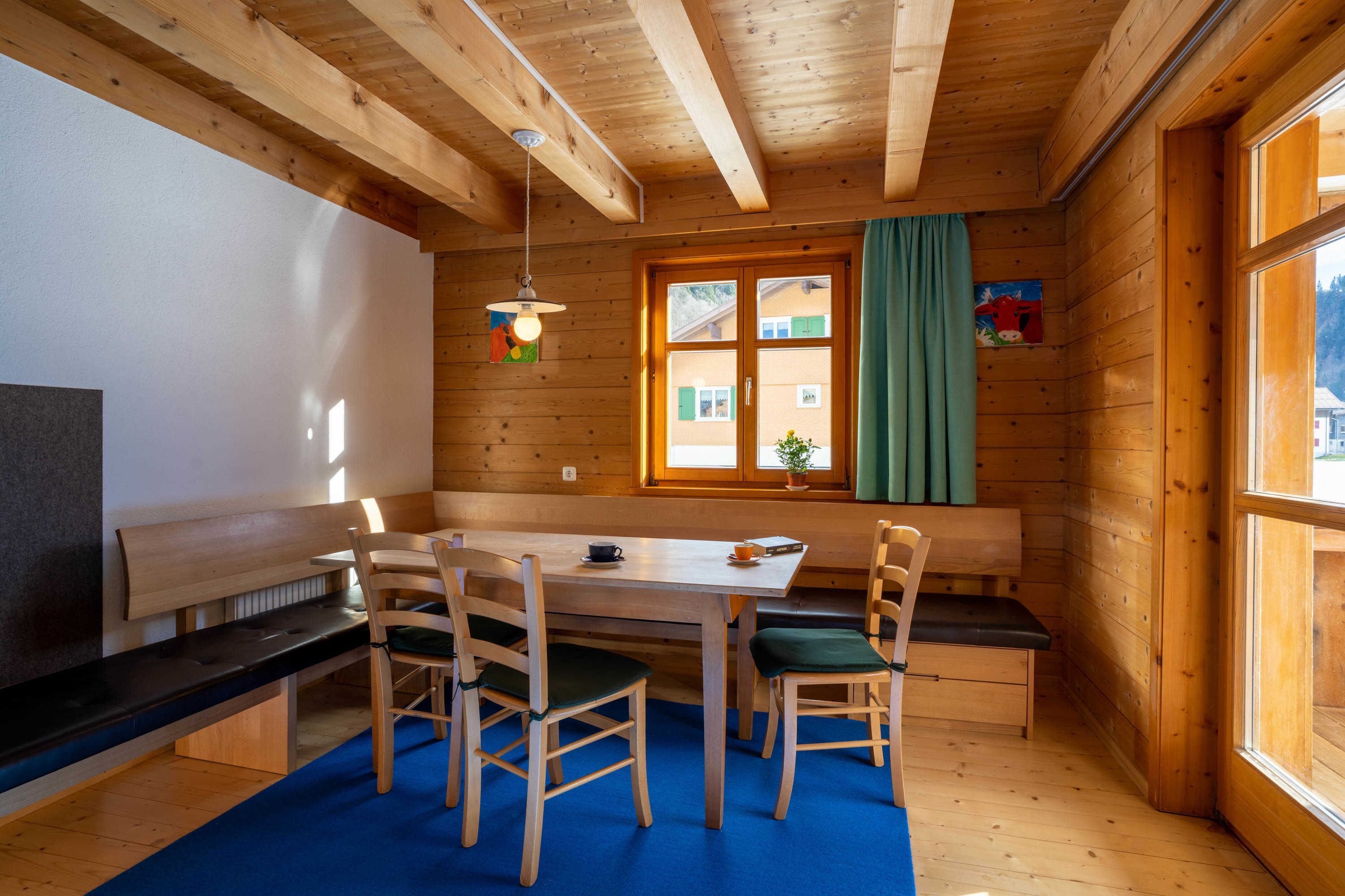 A wooden cabin interior with a dining table, chairs, a bench, a TV, and a window with curtains.