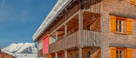 Un edificio de madera con balcón, paredes de ladrillo y una montaña nevada al fondo.