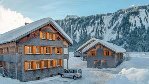 Eine Holzhütte mit steilem Dach, ein Auto davor und ein verschneiter Berg im Hintergrund.
