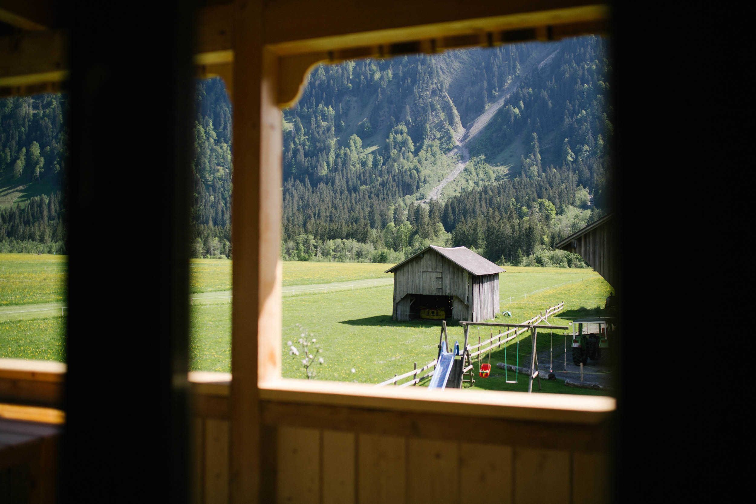 A wooden cabin with a view of a green field and a forested mountain range.