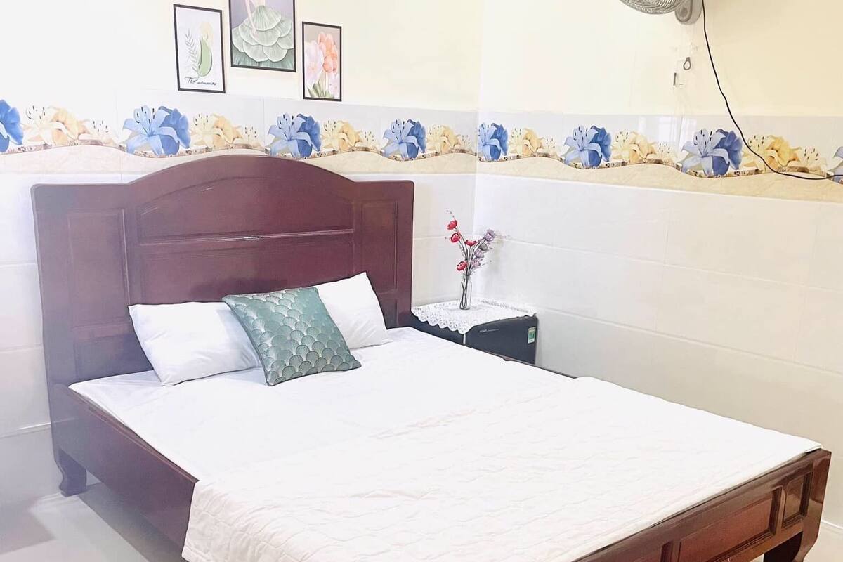 Una cama bien hecha con una colcha blanca, un cabecero de madera y una mesita de noche con una lámpara y un florero.