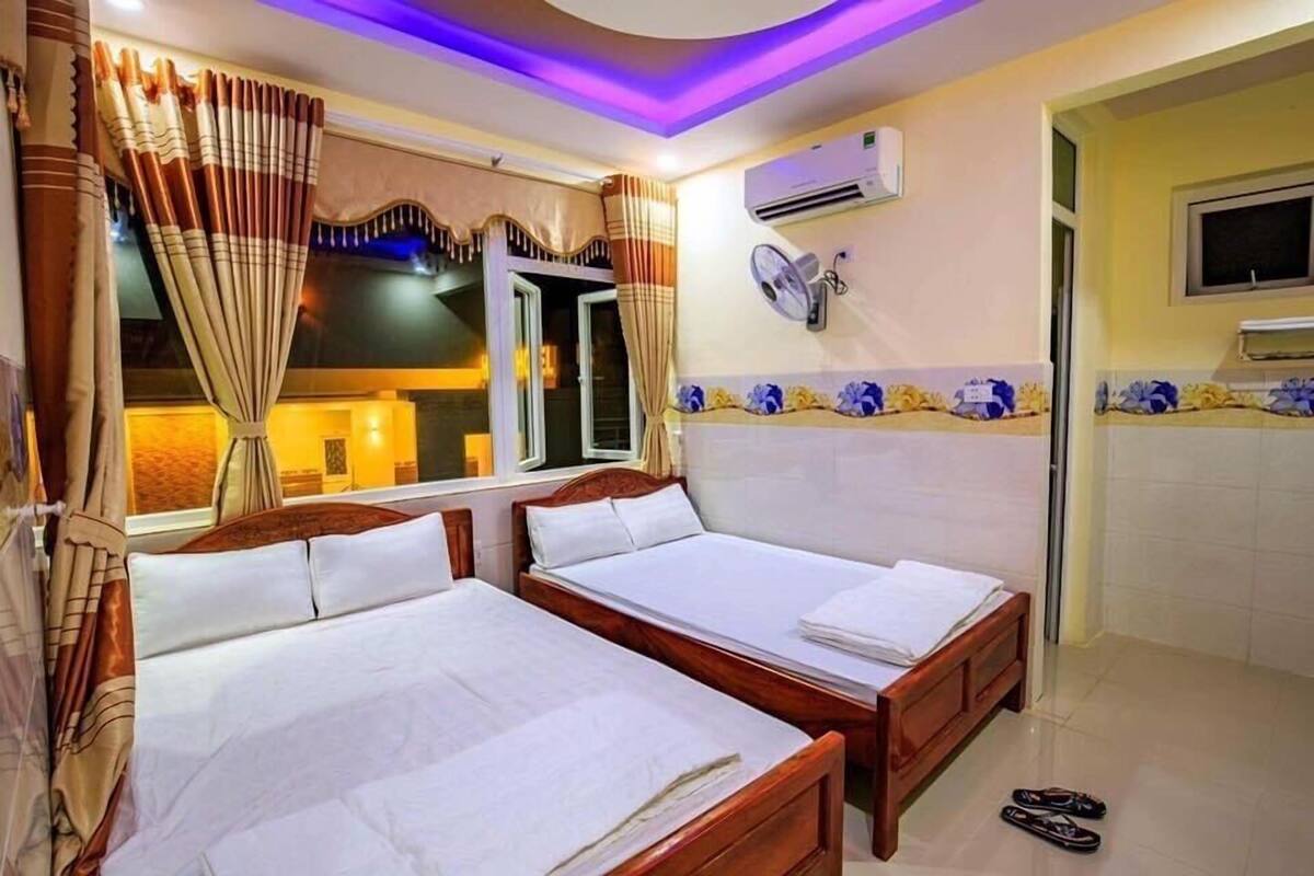 Habitación de hotel con dos camas de madera, una ventana con cortinas, un equipo de aire acondicionado y un ventilador de pared.