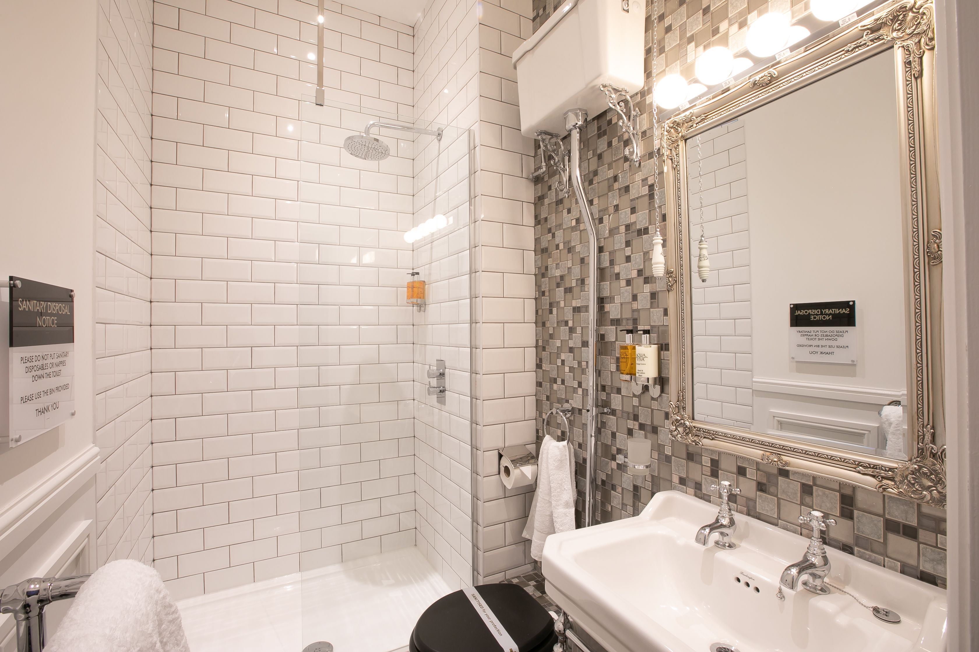 Un bagno con pareti piastrellate di bianco, una doccia con porta in vetro, un lavandino con due rubinetti e uno specchio con cornice decorata.