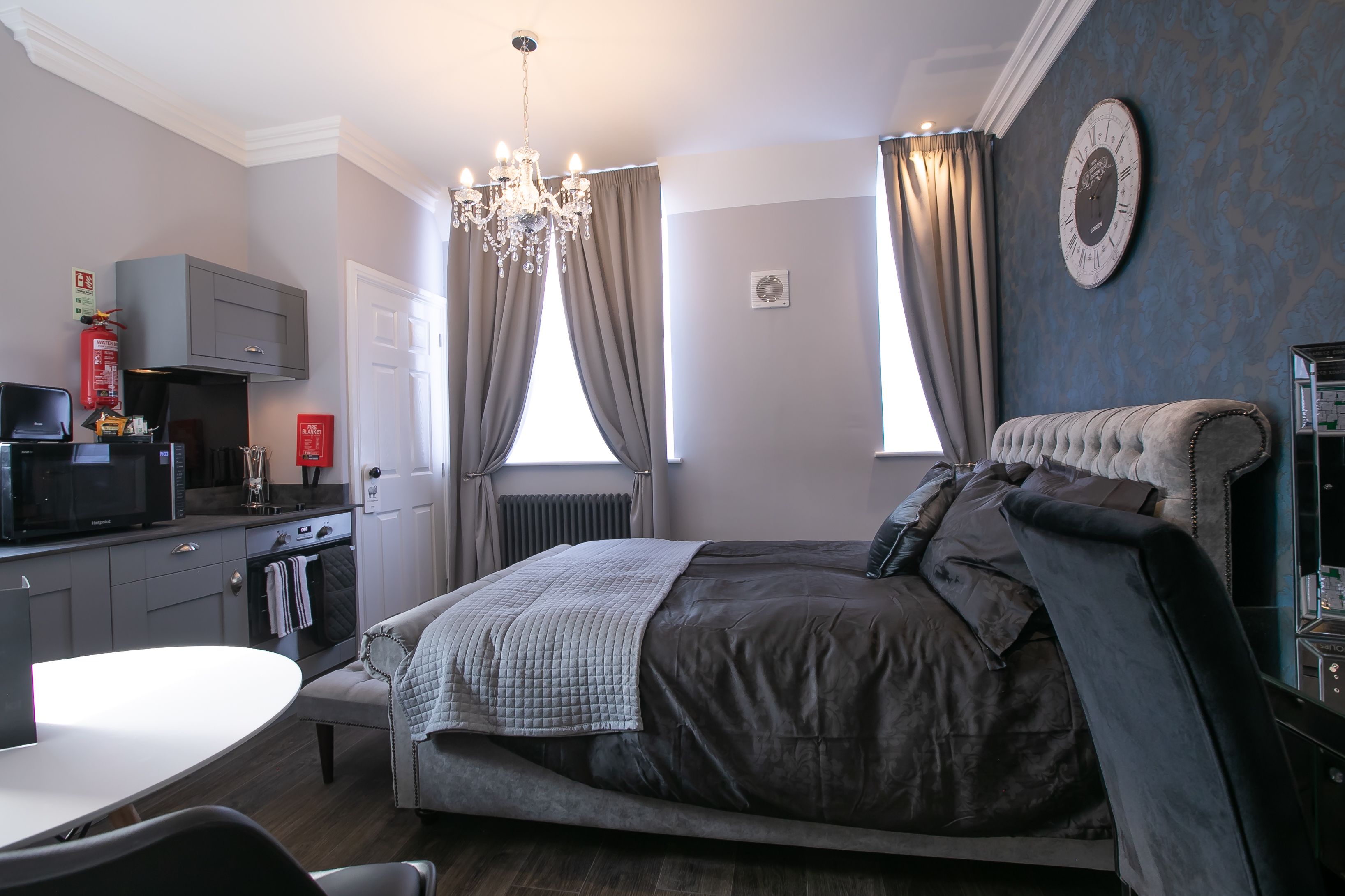 A bedroom with a bed, a chandelier, a clock, and a kitchen area.