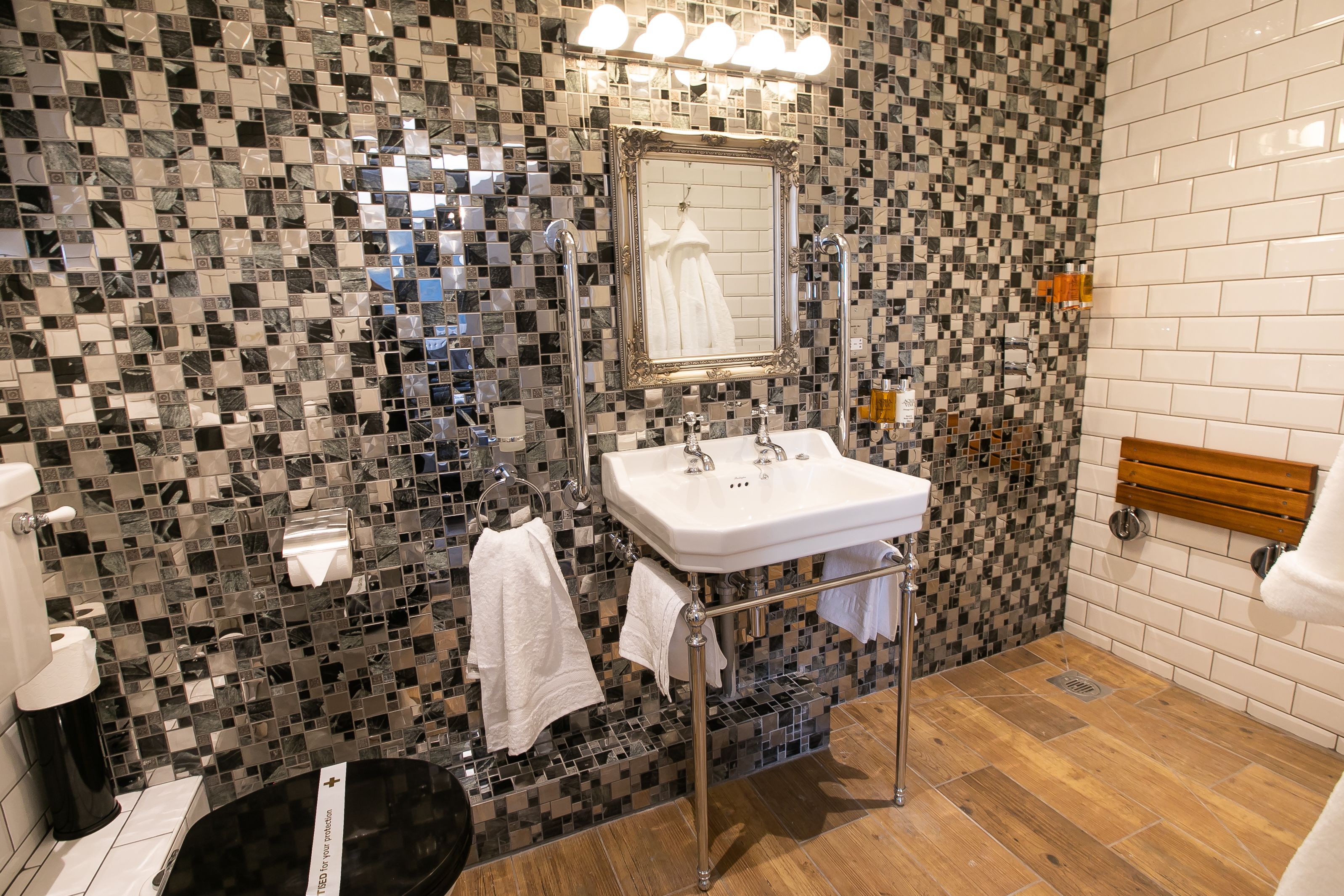 Un baño con pared de mosaicos, un lavabo blanco con dos grifos, un espejo y un banco de madera.