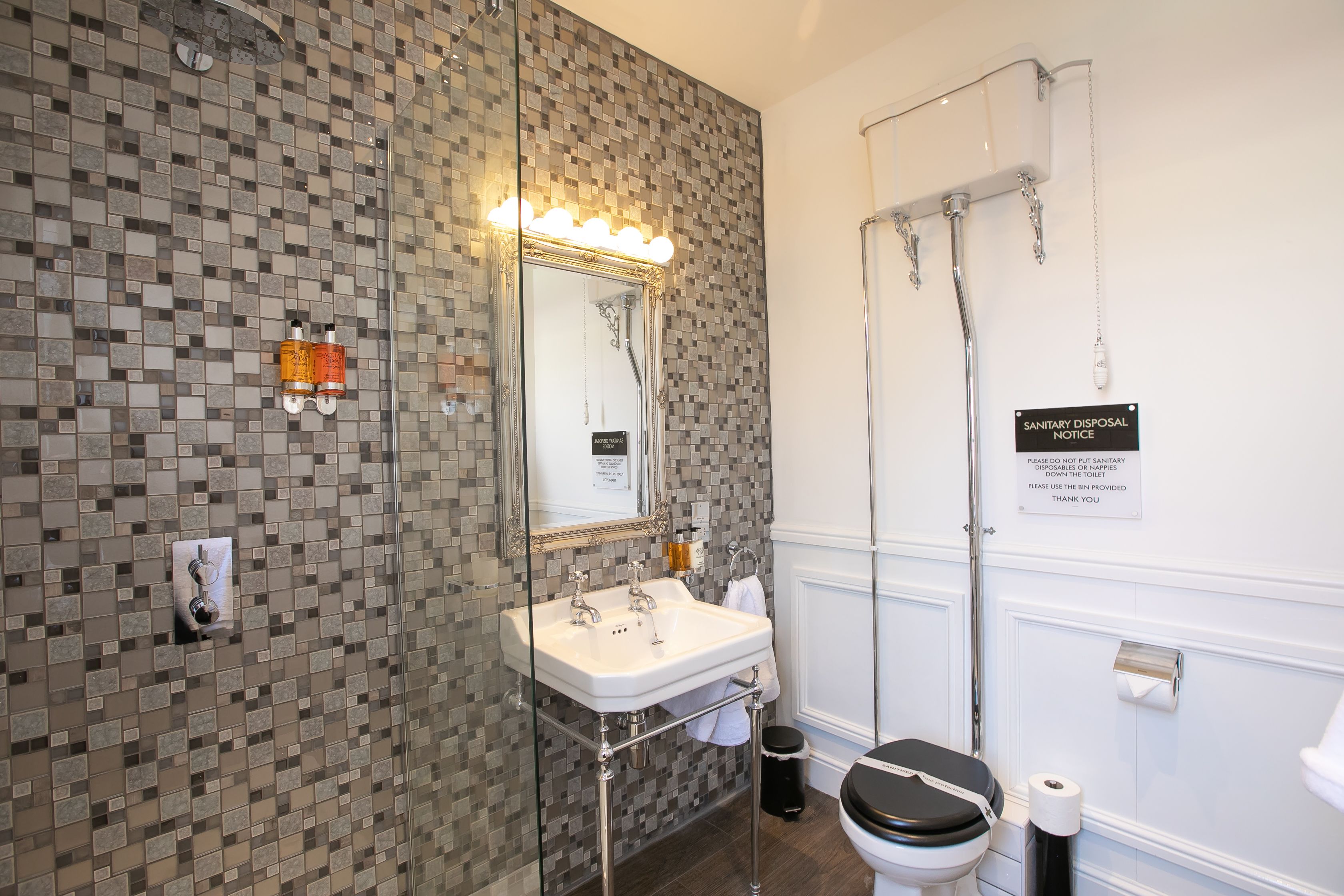 A bathroom with a tiled wall, a sink with a mirror, a toilet, and a shower area.