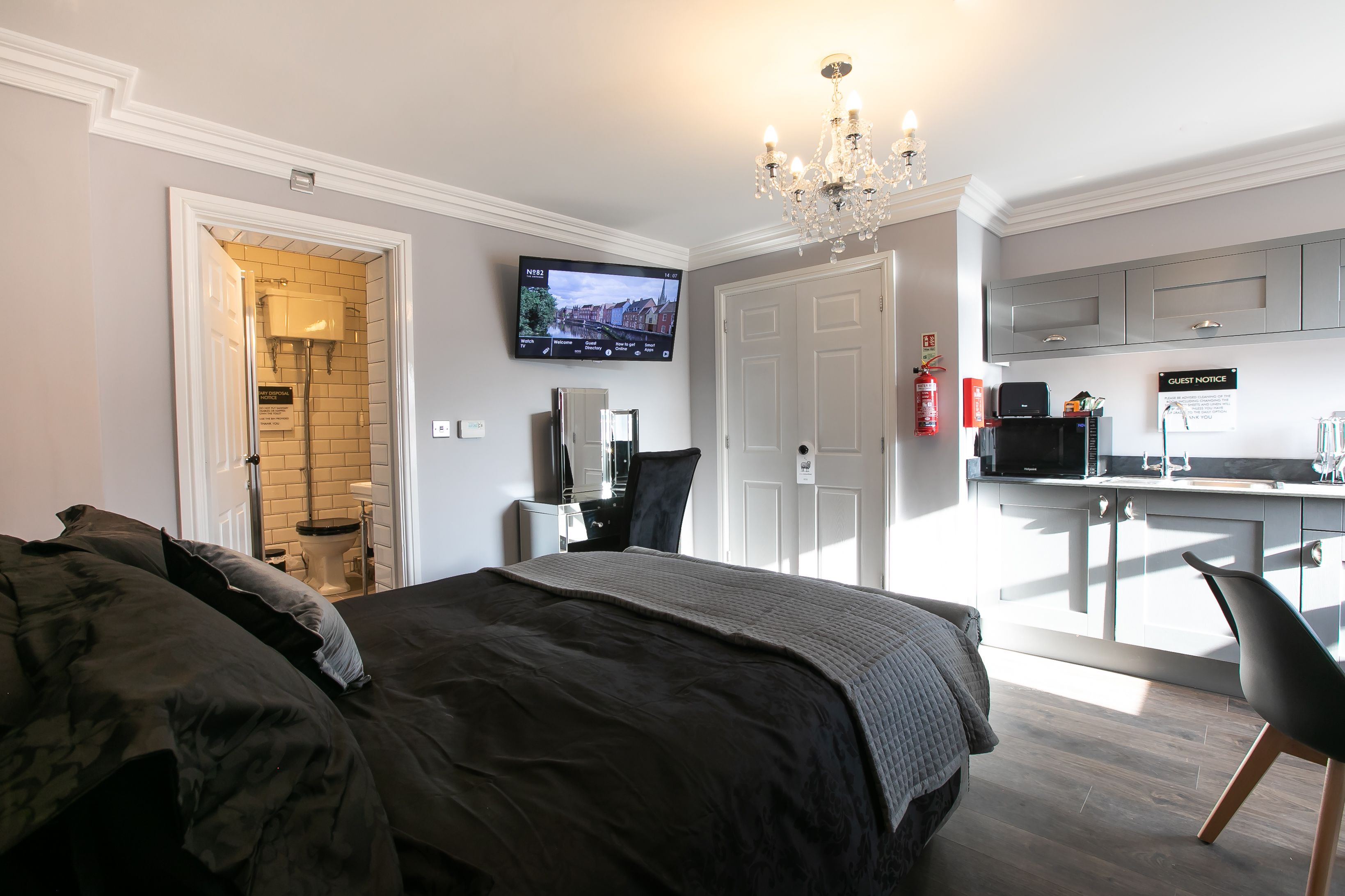 A hotel room with a bed, a TV mounted on the wall, a kitchenette, and a bathroom with a toilet and sink.