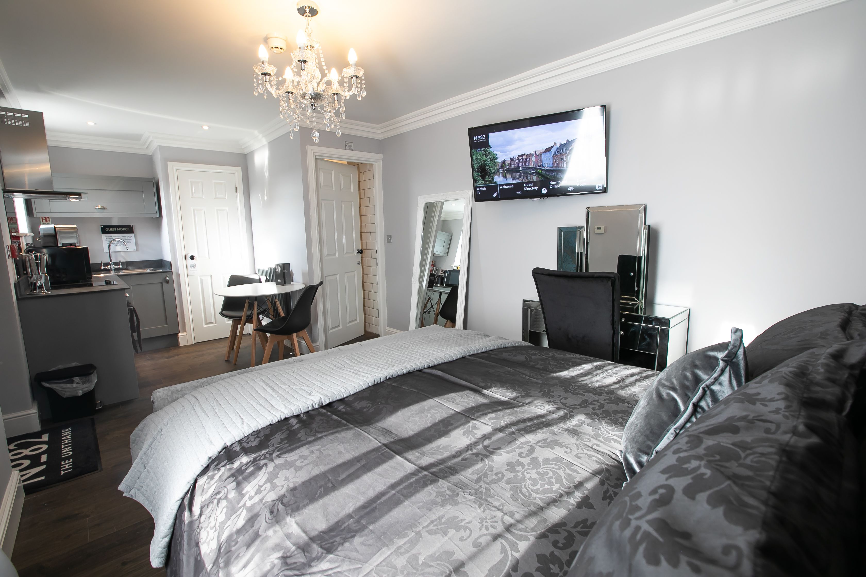 A modern hotel room with a large bed, a TV mounted on the wall, a desk with a chair, and a kitchen area in the background.