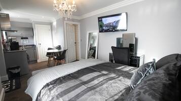 A modern hotel room with a large bed, a TV mounted on the wall, a desk with a chair, and a kitchen area in the background.