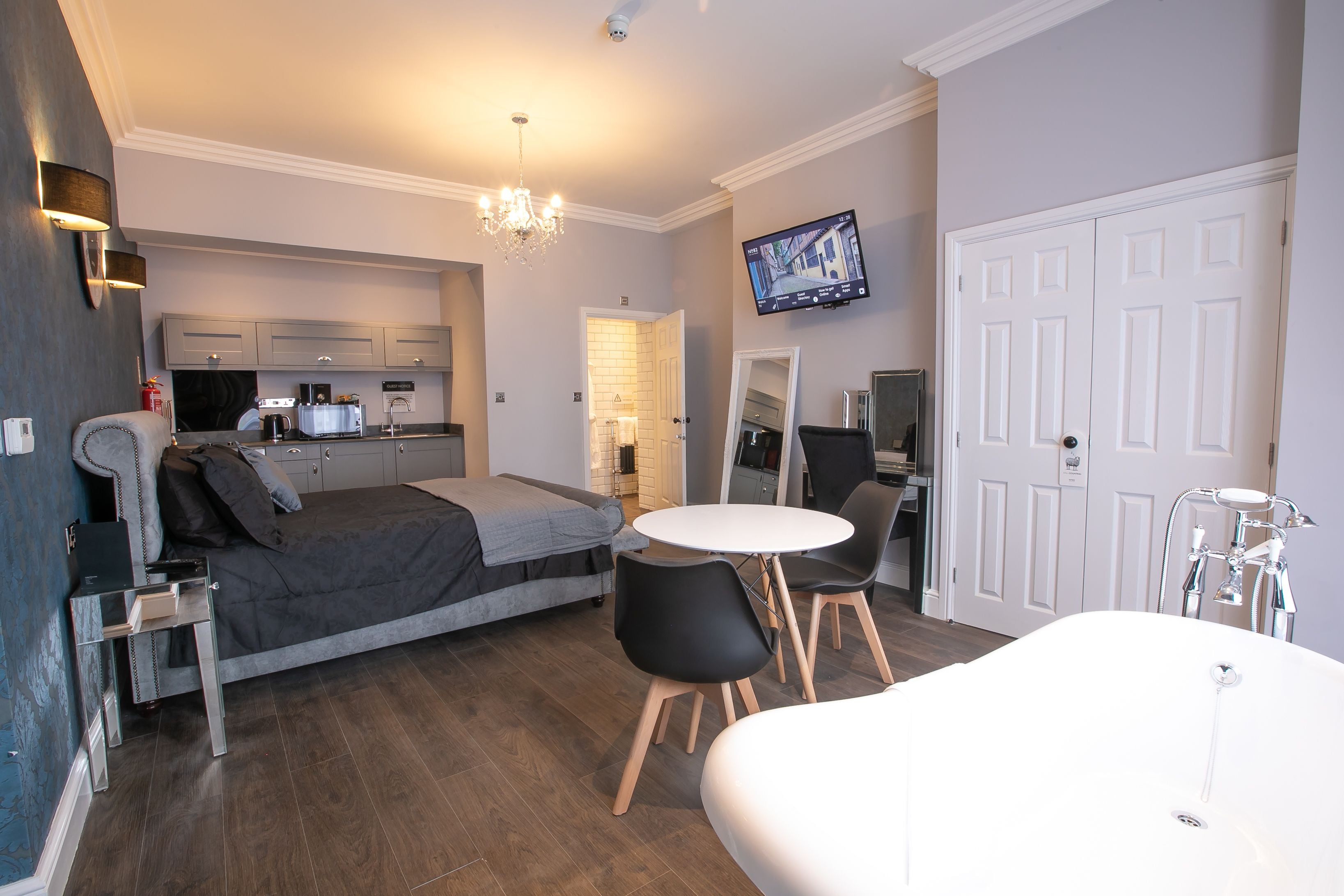 Een moderne slaapkamer met een bed, nachtkastje en een klein bureau. Er is een witte badkuip, een ronde eettafel met stoelen, en een aan de muur bevestigde televisie.