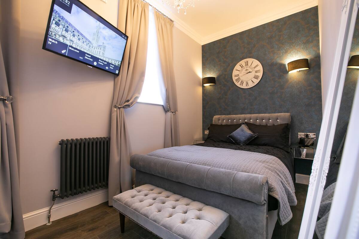 Un dormitorio moderno con una cama grande, un banco, un televisor montado en la pared y un reloj.