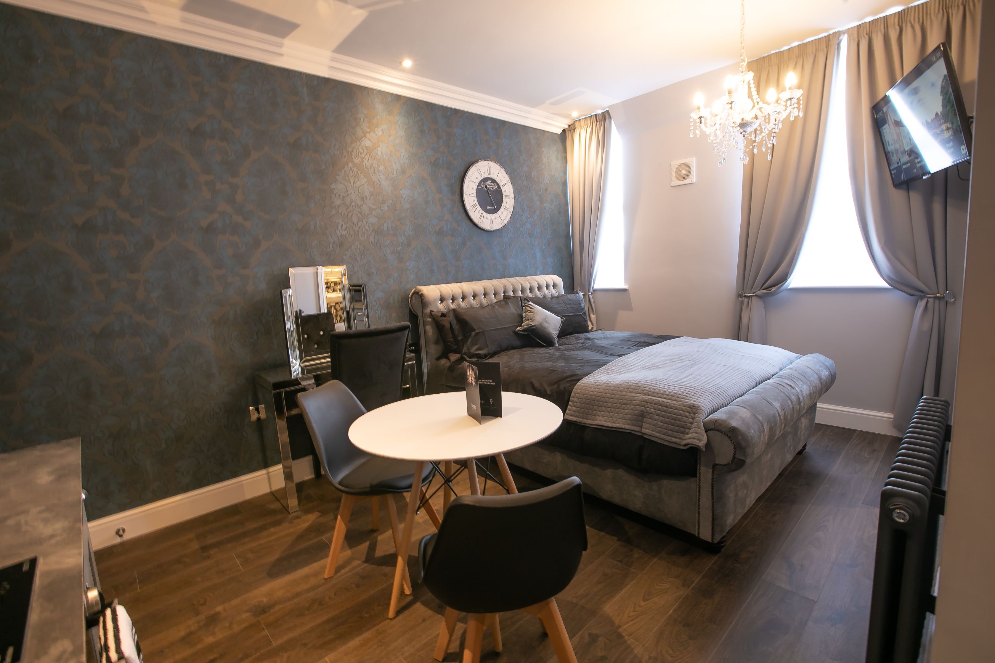 A bedroom with a bed, a desk with a chair, a round table, a television mounted on the wall, and a chandelier.