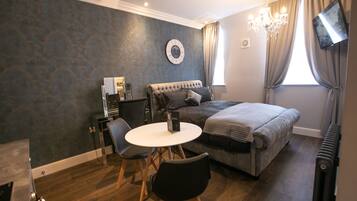 A bedroom with a bed, a desk with a chair, a round table, a television mounted on the wall, and a chandelier.
