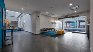 A modern hotel lobby with a seating area, a reception desk, and a large window offering a view of the street.