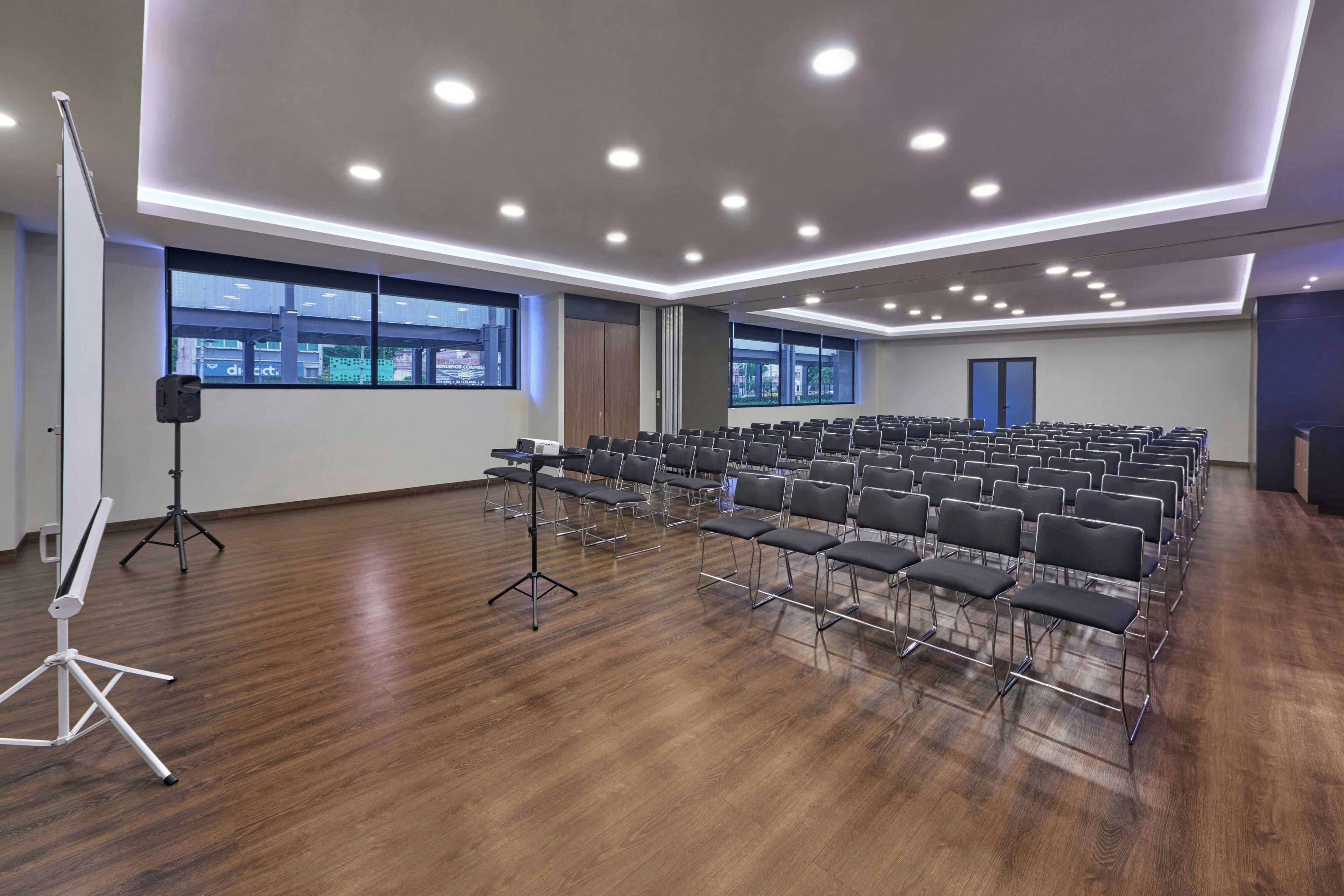 A conference room with rows of chairs, a projector stand, and large windows with city views.