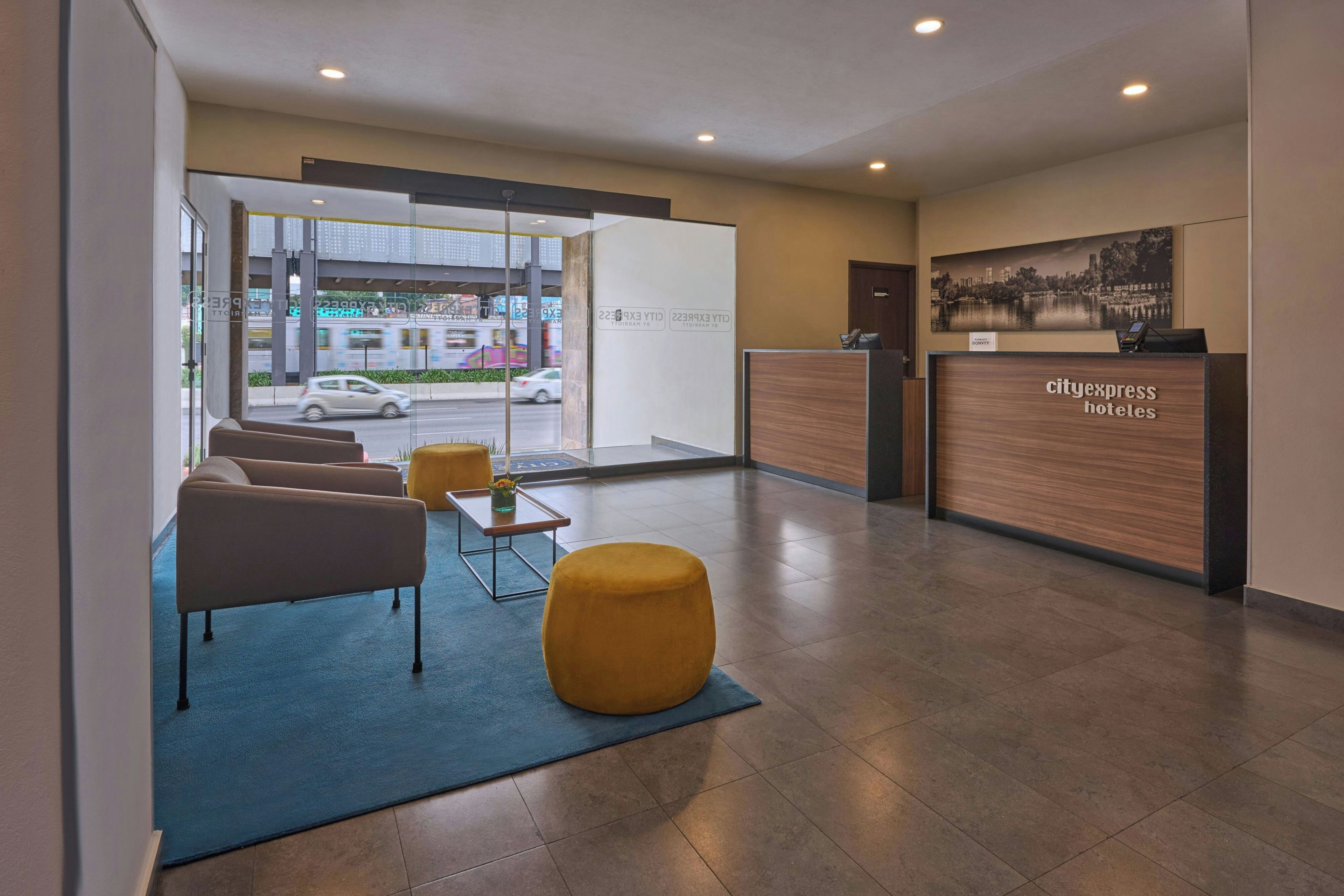 A hotel reception area with a reception desk, seating area with chairs and ottomans, and a view of the street through the glass door.