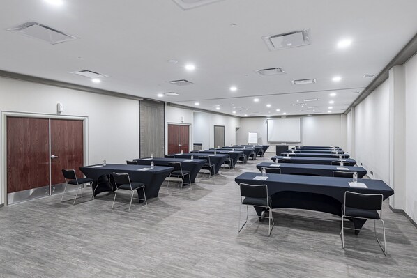 Une salle de conférence dotée de plusieurs tables et chaises, équipée de bouteilles d’eau et de documents, et un tableau blanc en arrière-plan.