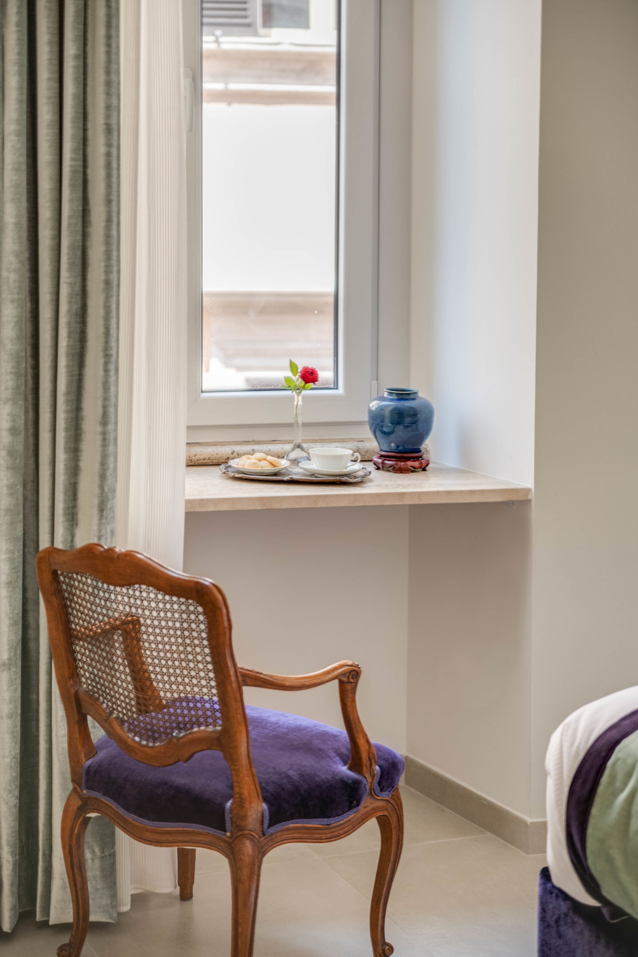 A room with a wooden chair with a purple cushion, a small table with a blue vase and a white cup, and a window with curtains.
