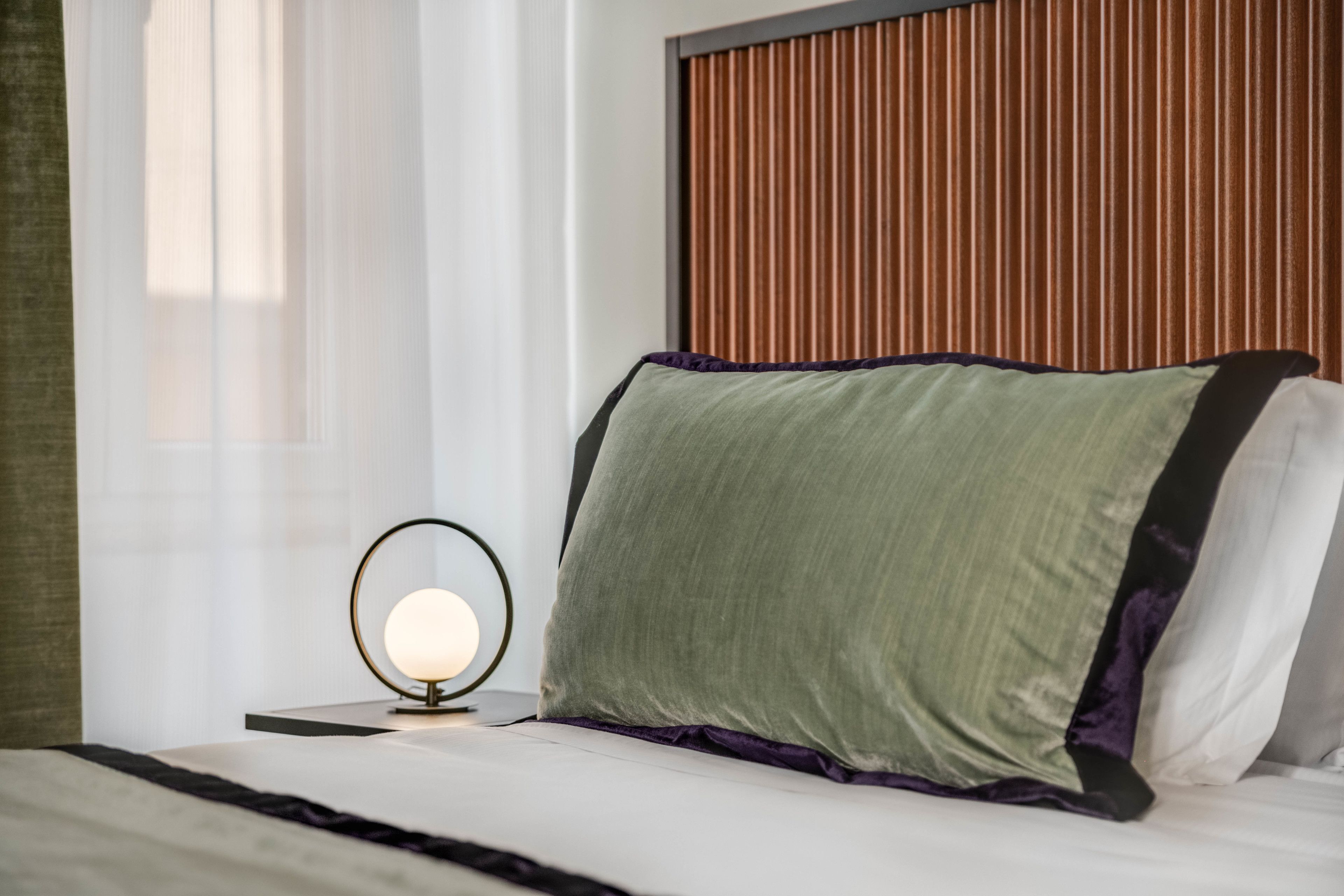 Un lit bien fait, avec un oreiller vert et un oreiller blanc, une table ronde avec une lampe et une tête de lit en bois.
