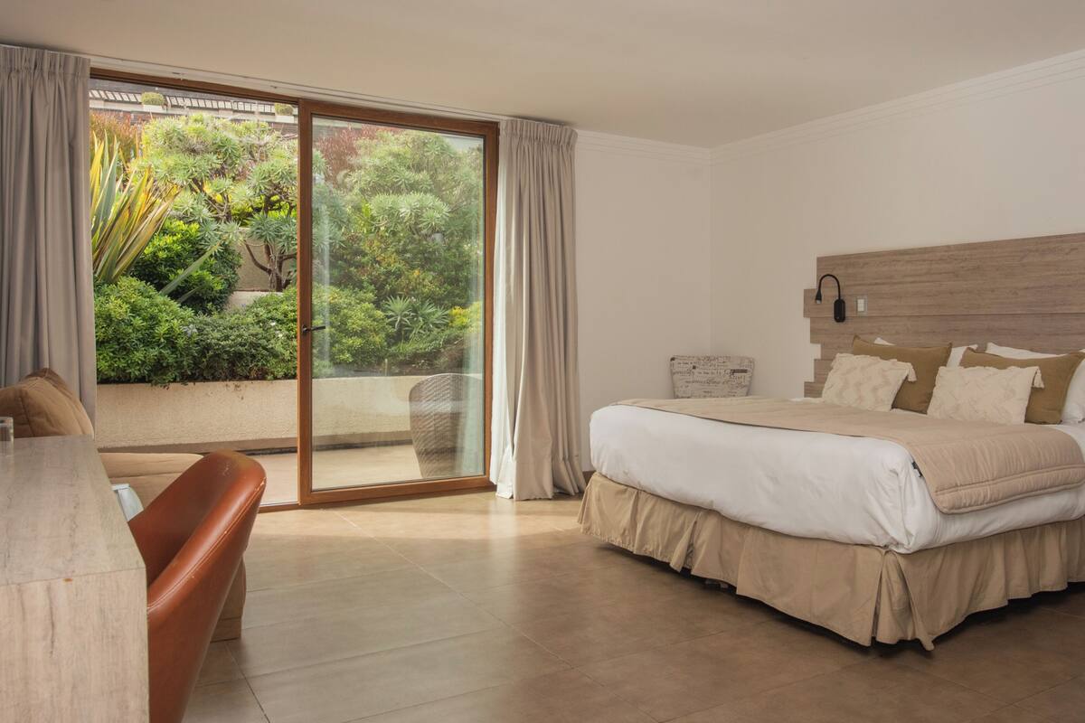 Un dormitorio con cama, escritorio, silla, mesita de noche y puerta corrediza de vidrio que da a un jardín.