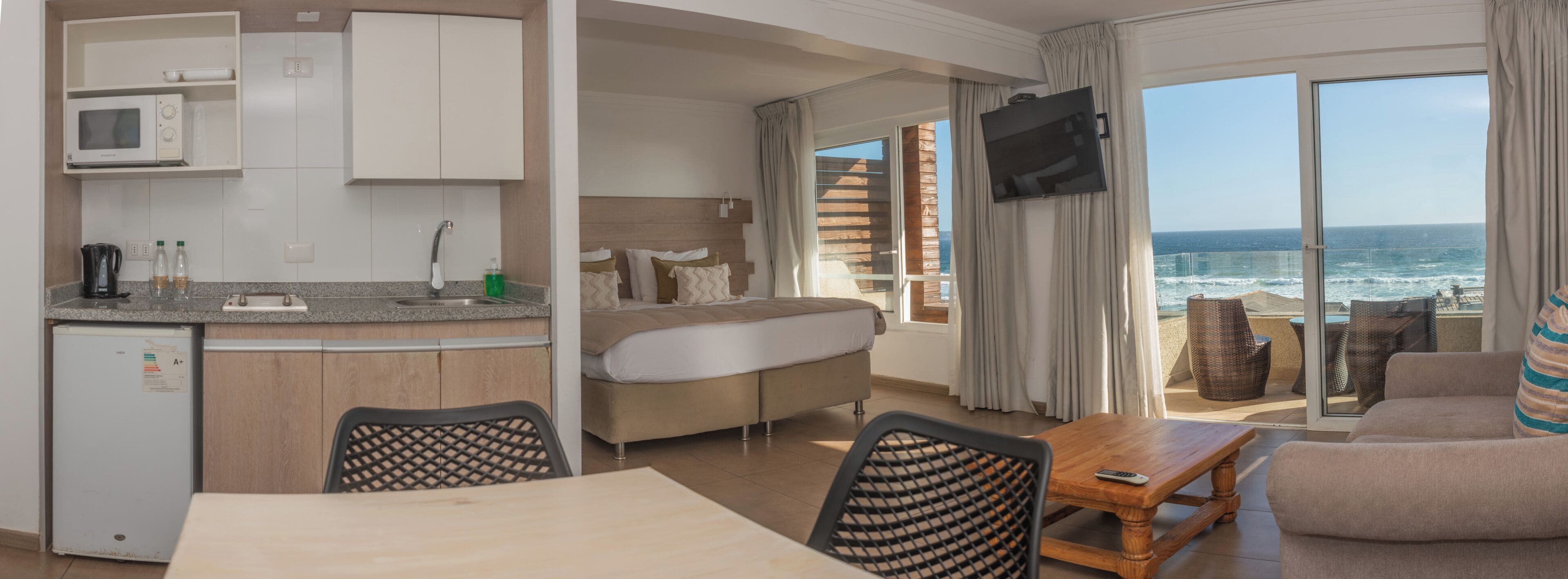 A compact hotel room with a kitchenette, a sofa, a dining table, and a TV.