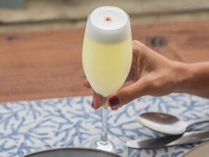 A hand holding a glass of yellow beverage, possibly a cocktail, with a frothy top. - Hotel Mae (Puchuncavi)
