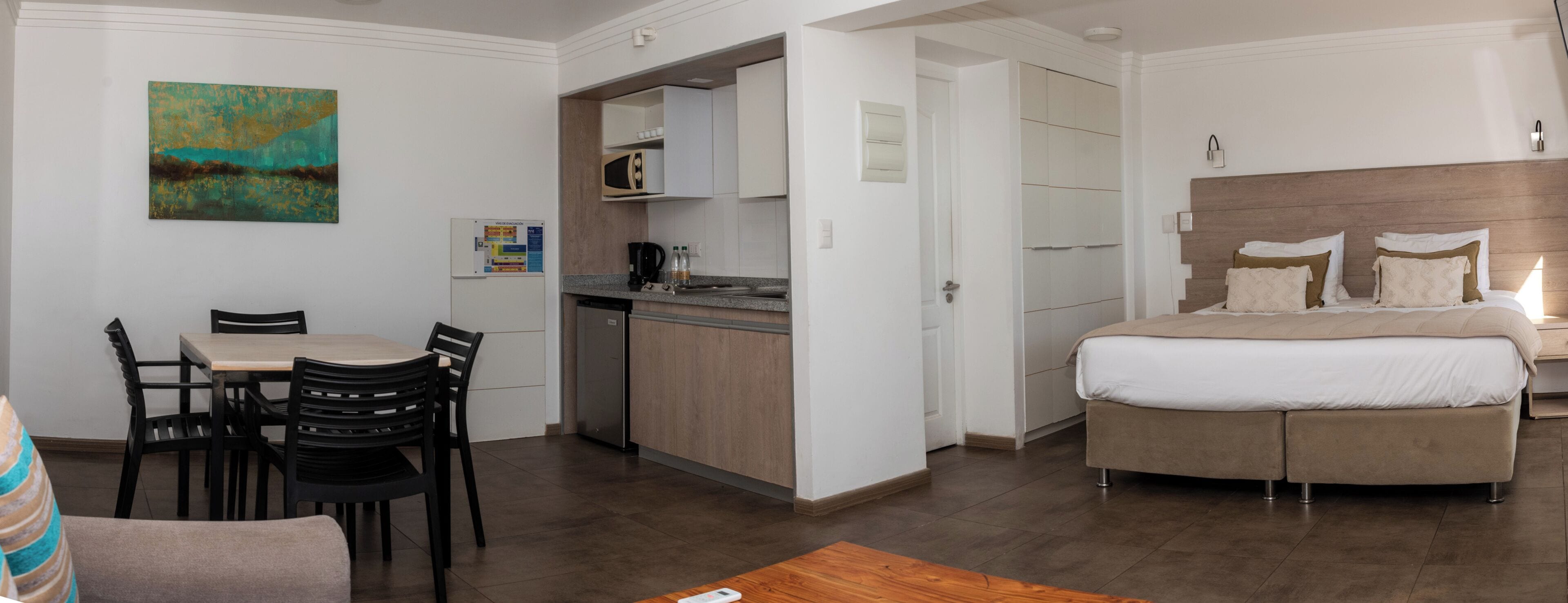 A hotel room with a bed, a dining table with chairs, a kitchenette, and a painting on the wall.
