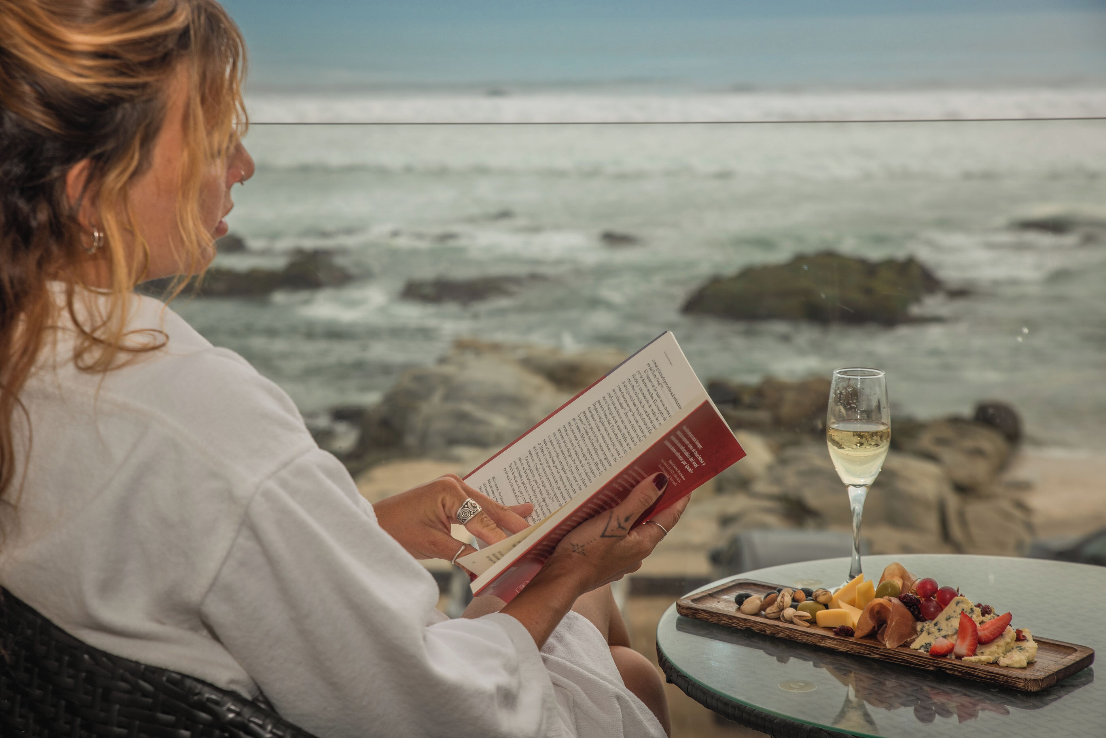 A person reading a book by a table with a glass of white wine and a charcuterie board.