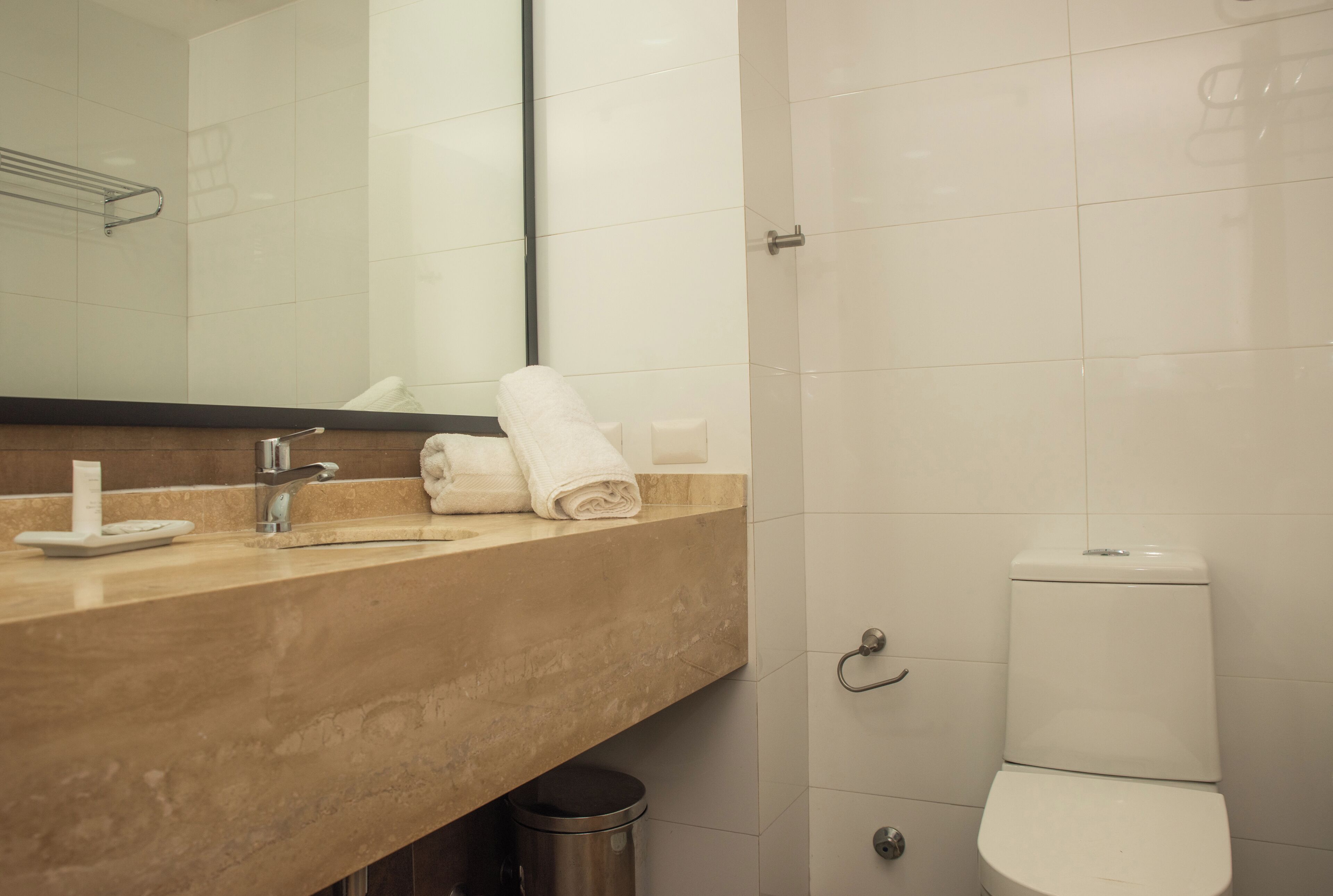 A bathroom with a beige countertop, a white sink, a mirror, a white toilet, and a trash can.