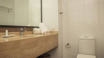 A bathroom with a beige countertop, a white sink, a mirror, a white toilet, and a trash can.